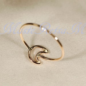 May include: A delicate gold-colored ring featuring a crescent moon design. The ring has a slender band and a detailed moon shape, creating a minimalist and elegant aesthetic. The ring is likely made of gold-toned metal.