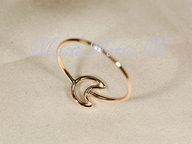 May include: A delicate gold-colored ring featuring a crescent moon design. The ring has a slender band and a detailed moon shape, creating a minimalist and elegant aesthetic. The ring is likely made of gold-toned metal.