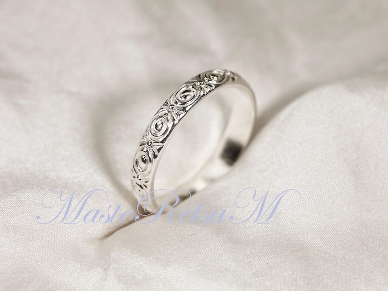 Puede incluir: Un anillo de plata con un intrincado dise&ntilde;o floral. El dise&ntilde;o del anillo presenta motivos circulares y florales repetidos, creando una superficie texturizada. El anillo se muestra sobre una tela blanca texturizada.