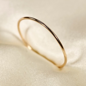 Puede incluir: Un delicado y fino anillo de oro. El anillo es una banda simple con un acabado pulido, sobre un fondo de tela suave blanquecina. El diseño es minimalista y elegante, adecuado para el uso diario o como anillo apilable. El diámetro del anillo es de aproximadamente 1,6 cm.