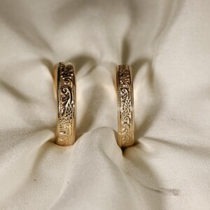 May include: Two gold-toned rings standing upright, each featuring an intricate, embossed floral and scrollwork pattern covering the entire outer surface of the band. The rings have a polished metallic finish and a consistent, rounded profile.