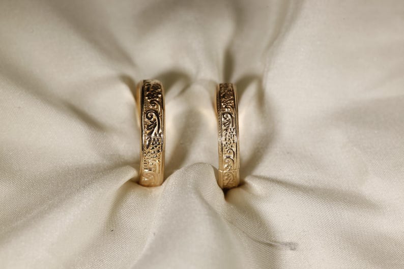 May include: Two gold-toned rings standing upright, each featuring an intricate, embossed floral and scrollwork pattern covering the entire outer surface of the band. The rings have a polished metallic finish and a consistent, rounded profile.