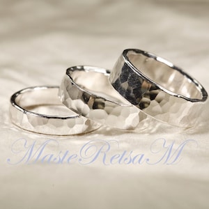 May include: Three silver rings with a hammered texture are displayed on a soft, off-white surface. The rings are close together, showcasing their detailed, textured surfaces. The rings are likely wedding bands or fashion accessories.