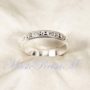 May include: A silver ring with an intricate, repeating pattern of floral and dot designs. The ring is set against a soft, off-white background. The ring's design is detailed and appears to be finely crafted. The text "Maste Retsa M" is in the foreground.