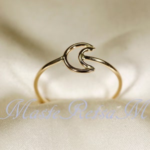May include: A delicate gold-colored ring featuring a crescent moon design. The ring has a thin band and a minimalist style. The ring is shown on a white satin surface. The text "Maste Reisa M" is at the bottom of the image.