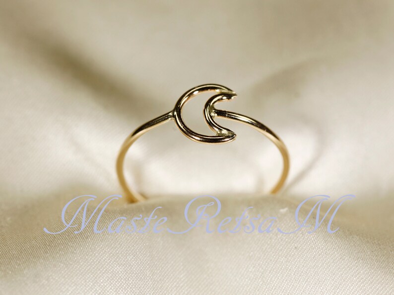 May include: A delicate gold-colored ring featuring a crescent moon design. The ring has a thin band and a minimalist style. The ring is shown on a white satin surface. The text "Maste Reisa M" is at the bottom of the image.