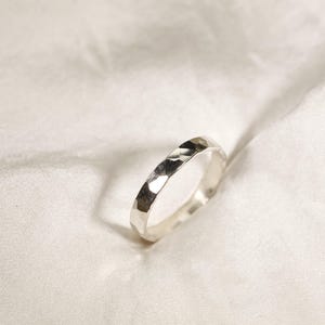 May include: A hammered silver ring with a textured surface. The ring is set against a white, textured background. The ring is a simple band design, reflecting light.