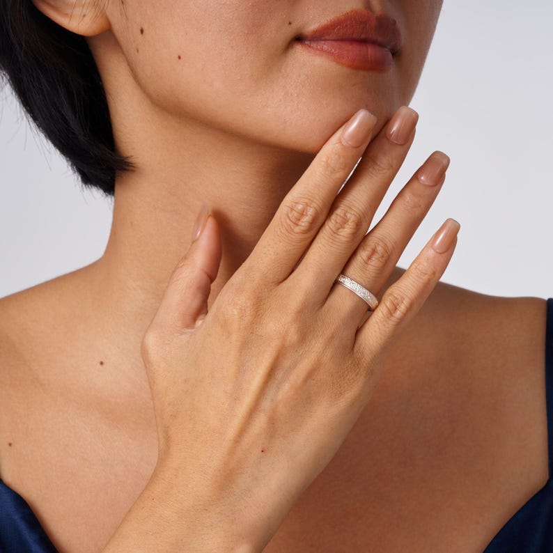 May include: A silver-toned band ring featuring a textured, sparkling surface. The ring is worn on the ring finger, showcasing a slim, uniform width and a metallic finish that reflects light across its entire circumference.