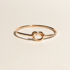 May include: A delicate rose gold ring featuring a knot design. The ring is set against a soft, neutral background, highlighting the polished metal and the ring's simple elegance. A minimalist piece of jewelry.