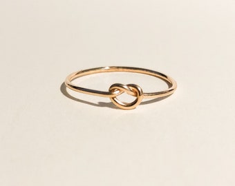 May include: A rose gold ring with a knot design. The ring is shown against a neutral background, highlighting the polished metal and the ring's simple elegance. A minimalist piece of jewellery.