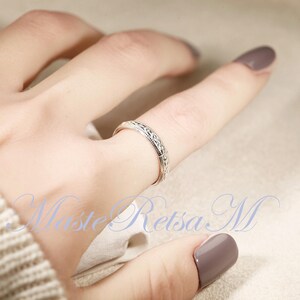 May include: A silver ring with an intricate, repeating pattern encircles a finger. The ring's design features small, detailed elements. The image is well-lit, highlighting the ring's texture and the subtle details of the design. The ring is a piece of jewelry.