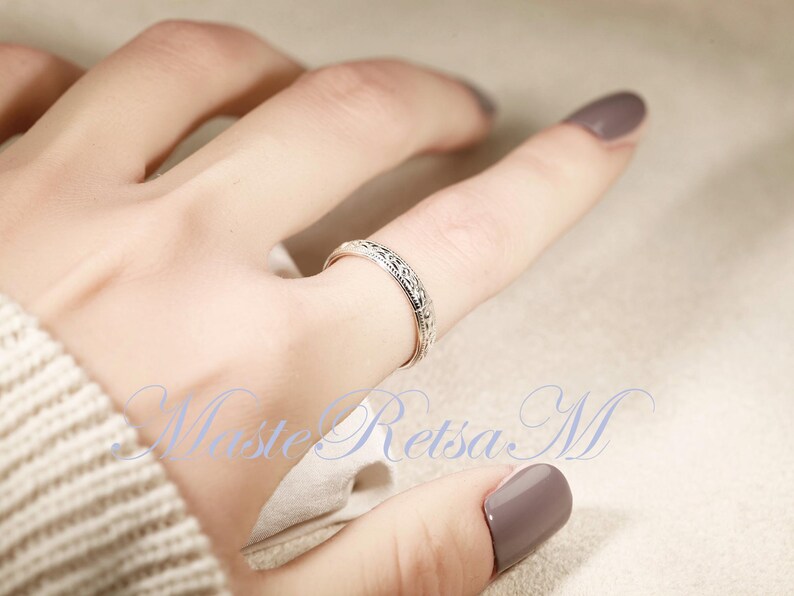 May include: A silver ring with an intricate, repeating pattern encircles a finger. The ring's design features small, detailed elements. The image is well-lit, highlighting the ring's texture and the subtle details of the design. The ring is a piece of jewelry.