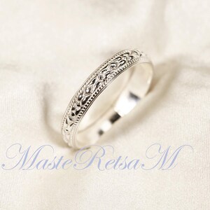 May include: A silver ring with an intricate, repeating pattern of diamond and floral shapes. The ring is set against a white, textured background. The ring's design is detailed and elegant.