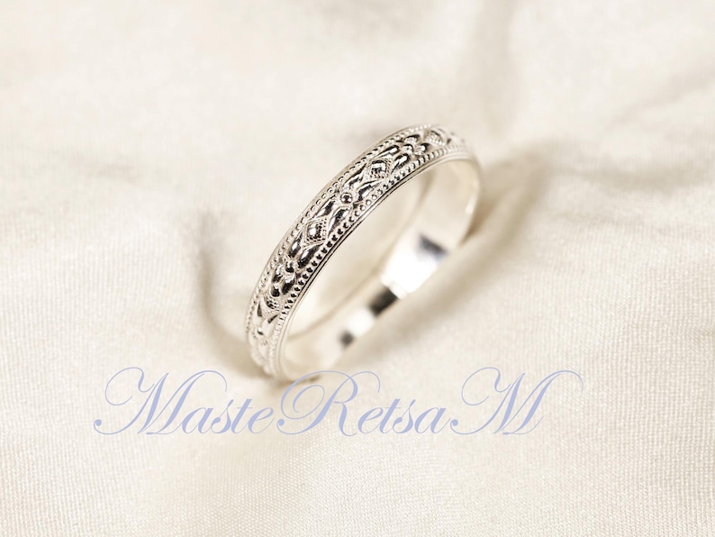 May include: A silver ring with an intricate, repeating pattern of diamond and floral shapes. The ring is set against a white, textured background. The ring's design is detailed and elegant.