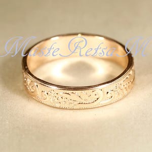 May include: A rose gold band ring featuring an intricate engraved floral pattern along the outer surface. The metal has a polished finish and the text Maste Refsa M is overlaid in light blue script across the center of the ring's circular opening.