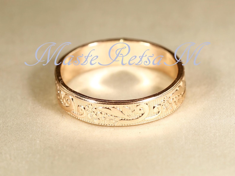May include: A rose gold band ring featuring an intricate engraved floral pattern along the outer surface. The metal has a polished finish and the text Maste Refsa M is overlaid in light blue script across the center of the ring's circular opening.