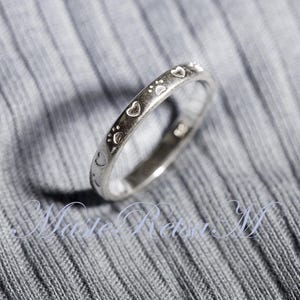 May include: A silver-toned metal ring featuring a repeating pattern of engraved hearts and paw prints around the outer band. The ring has a smooth, polished finish and is positioned at an angle to display the continuous design of the symbols.
