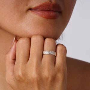 May include: A silver-toned metal ring featuring a continuous embossed floral pattern around the band. The ring has a polished finish and a flat profile, worn on the finger, showcasing intricate engraved details across the entire outer surface.