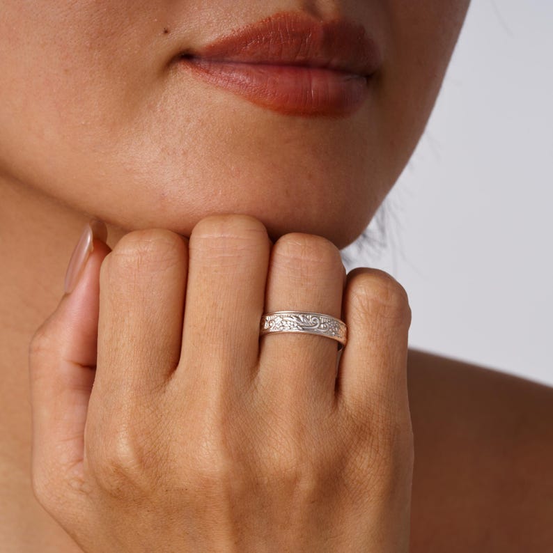 May include: A silver-toned metal ring featuring a continuous embossed floral pattern around the band. The ring has a polished finish and a flat profile, worn on the finger, showcasing intricate engraved details across the entire outer surface.