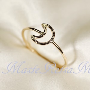 May include: A gold-colored ring featuring a crescent moon design. The ring has a delicate band and the moon shape is open, creating a minimalist aesthetic. The ring is displayed on a soft, light-colored surface.