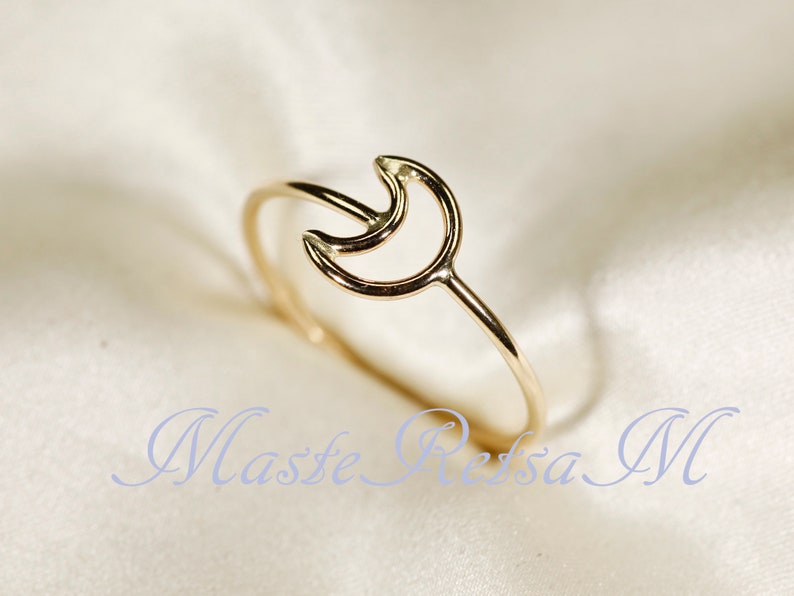 May include: A gold-colored ring featuring a crescent moon design. The ring has a delicate band and the moon shape is open, creating a minimalist aesthetic. The ring is displayed on a soft, light-colored surface.