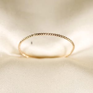 Può includere: Un delicato braccialetto color oro con un design strutturato e perlato. Il braccialetto è aperto e poggia su un tessuto color crema, mostrando la sua semplice eleganza. Il gioiello è un accessorio elegante.
