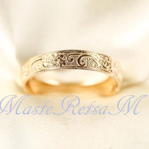 May include: A gold-toned metal ring featuring an intricate embossed floral and scrollwork pattern across its outer surface. The band has a smooth, polished interior and a consistent width. The text Maste Retsa M is visible in blue.