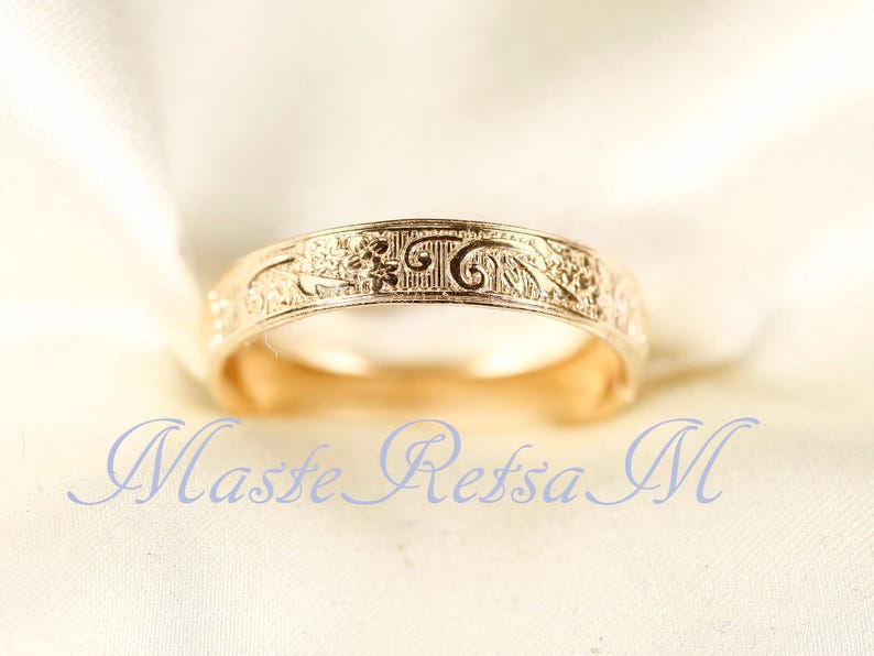 May include: A gold-toned metal ring featuring an intricate embossed floral and scrollwork pattern across its outer surface. The band has a smooth, polished interior and a consistent width. The text Maste Retsa M is visible in blue.