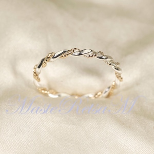 Puede incluir: Un anillo trenzado en tonos plateados y dorados. El anillo presenta un diseño trenzado con secciones alternas de plata y oro. El anillo está sobre un fondo suave y blanquecino.