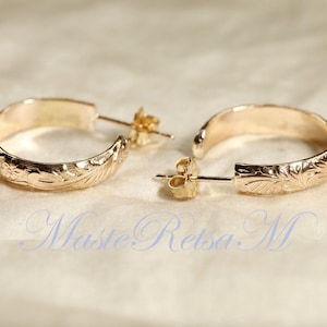 May include: A pair of gold-colored hoop earrings with a floral pattern. The earrings are half-hoop style with a post and butterfly backing. The earrings are displayed on a cream-colored surface. The text "Maste Retsa M" is visible in the image.