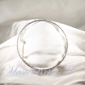 May include: A silver bangle bracelet with a textured design. The bracelet is lying on a white fabric surface.