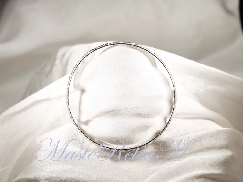 May include: A silver bangle bracelet with a textured design. The bracelet is lying on a white fabric surface.