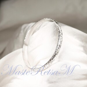May include: A silver bangle bracelet with an intricate floral design. The bracelet is on a white fabric background.