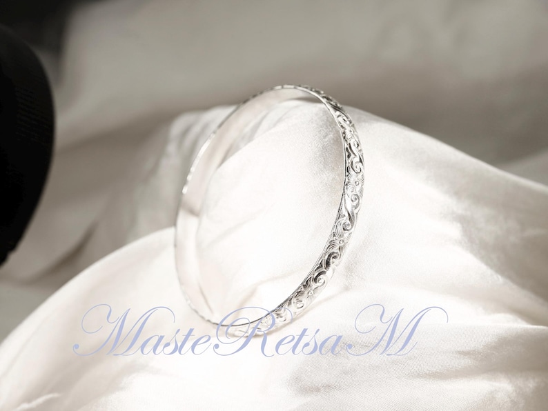 May include: A silver bangle bracelet with an intricate floral design. The bracelet is on a white fabric background.