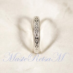 May include: A silver ring with an intricate, repeating floral pattern. The ring is set against a white background, and the design is visible around the entire band. The ring is a close-up shot, highlighting the details of the design.