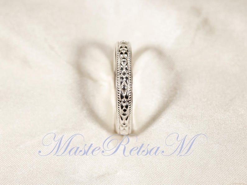 May include: A silver ring with an intricate, repeating floral pattern. The ring is set against a white background, and the design is visible around the entire band. The ring is a close-up shot, highlighting the details of the design.