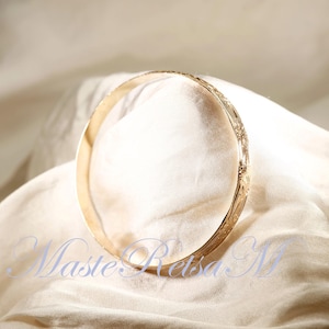 May include: A gold bangle bracelet with a delicate floral design. The bracelet is lying on a white fabric background.