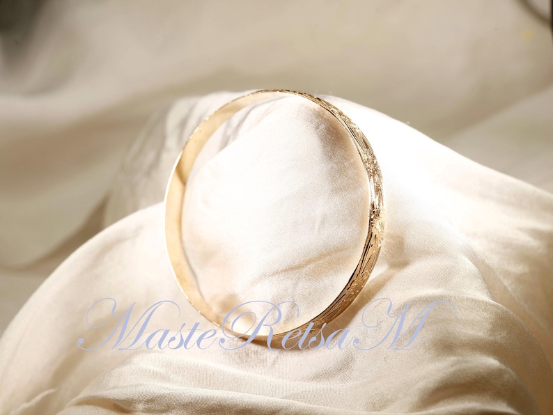 May include: A gold bangle bracelet with a delicate floral design. The bracelet is lying on a white fabric background.