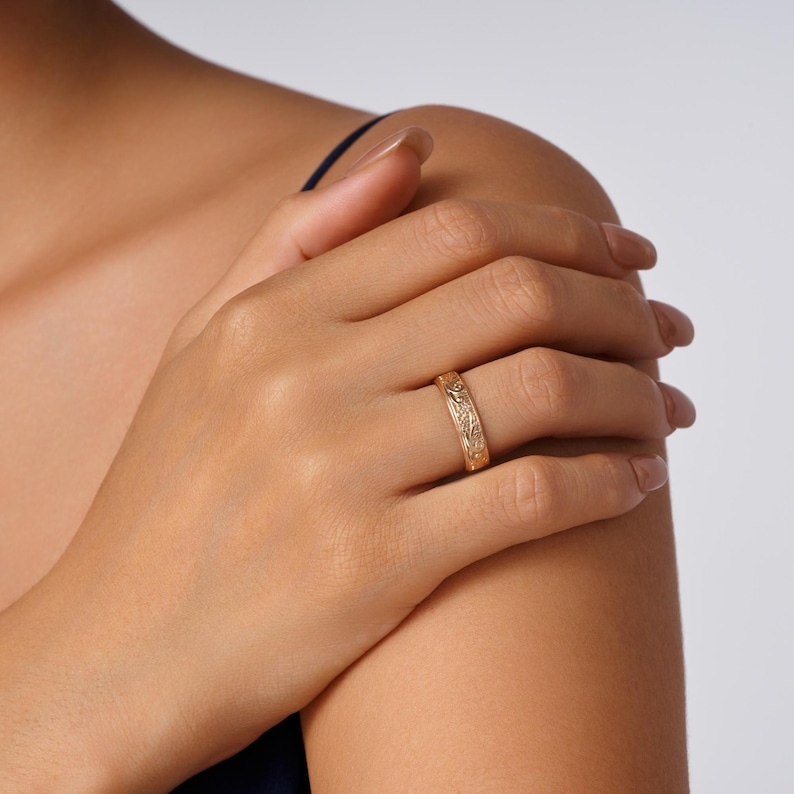 May include: A gold band ring featuring an intricate, embossed floral pattern that wraps around the entire circumference of the metal. The ring has a polished finish and is worn on the ring finger, showcasing detailed craftsmanship and texture.