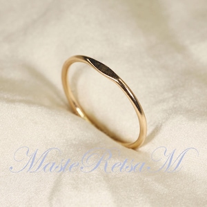 May include: A thin, polished gold ring featuring a small, flat, marquise-shaped focal point on the top of the band. The ring has a smooth, rounded shank and a reflective metallic finish. The text Maste Retsa M is visible in blue script.