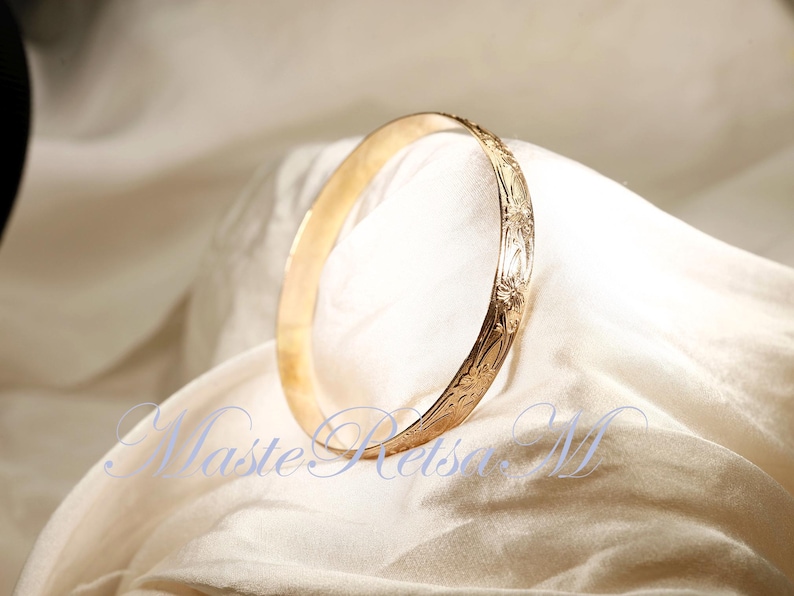 May include: A gold bangle bracelet with an intricate floral design. The bracelet is smooth and shiny, and the floral pattern is raised and detailed.