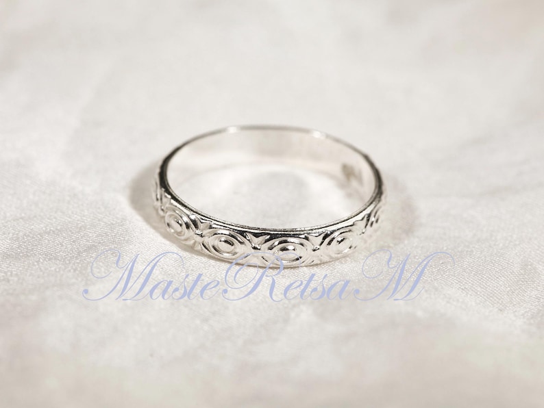 Puede incluir: Un anillo de plata con un patr&oacute;n circular repetido alrededor de la banda. El anillo est&aacute; sobre un fondo blanco texturizado. Es una pieza de joyer&iacute;a.
