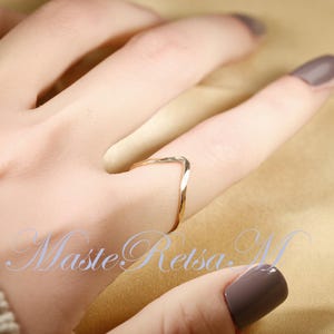 May include: A gold-toned ring with a twisted, V-shaped design. The ring is worn on a finger with light brown nail polish.