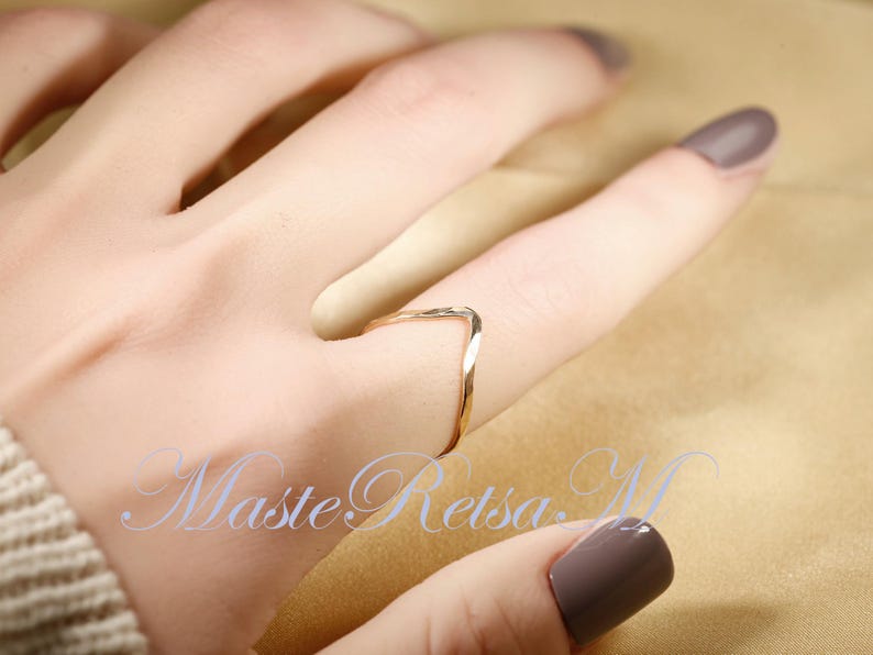 May include: A gold-toned ring with a twisted, V-shaped design. The ring is worn on a finger with light brown nail polish.