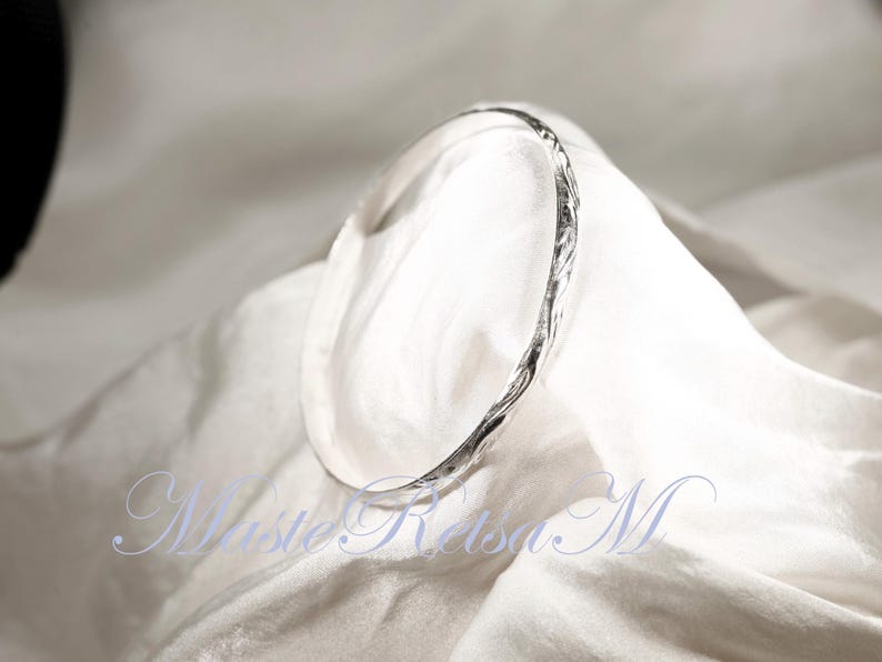 May include: A silver bangle bracelet with a textured, twisted design. The bracelet is on a white fabric background.
