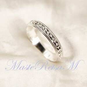 May include: A silver ring with an intricate, repeating pattern of dots and floral designs. The ring is set against a white background, highlighting its detailed craftsmanship. The ring's design is symmetrical and elegant.