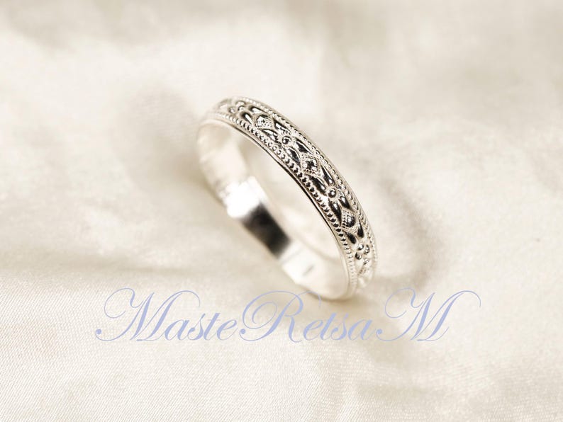 May include: A silver ring with an intricate, repeating pattern of dots and floral designs. The ring is set against a white background, highlighting its detailed craftsmanship. The ring's design is symmetrical and elegant.