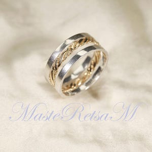 Könnte beinhalten: Zweifarbiger Ring mit einem silbernen Band und einer goldfarbenen geflochtenen Mitte. Der Ring hat eine polierte Oberfläche und ein strukturiertes Design. Der Ring befindet sich auf weißem Hintergrund. Der Text "Maste Retsa M" befindet sich unten im Bild.