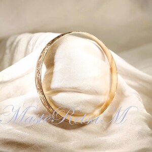 May include: A gold bangle bracelet with an intricate engraved design. The bracelet is lying on a white fabric surface.