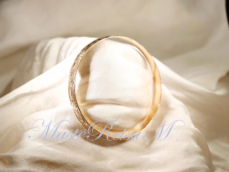 May include: A gold bangle bracelet with an intricate engraved design. The bracelet is lying on a white fabric surface.